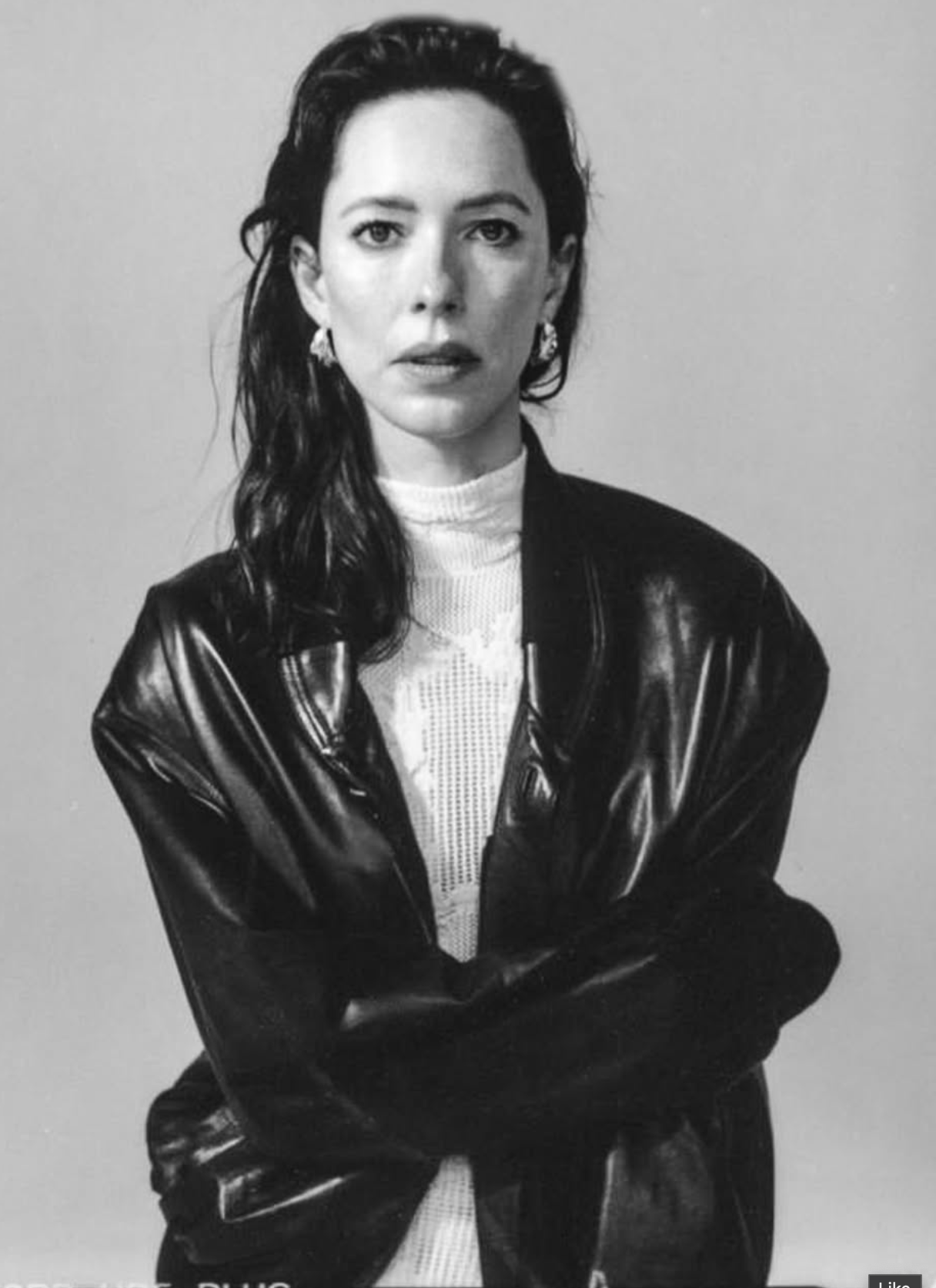 A black and white portrait of a woman with dark hair wearing a shiny black leather jacket over a white ribbed turtleneck, posed against a plain light gray background.