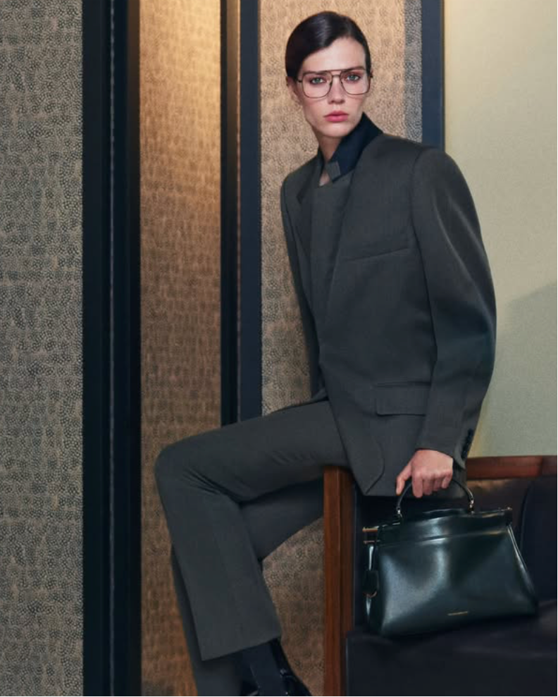 A model wearing a dark charcoal gray tailored suit, black shirt, and black patent leather shoes sits in a wooden armchair holding a black leather handbag.