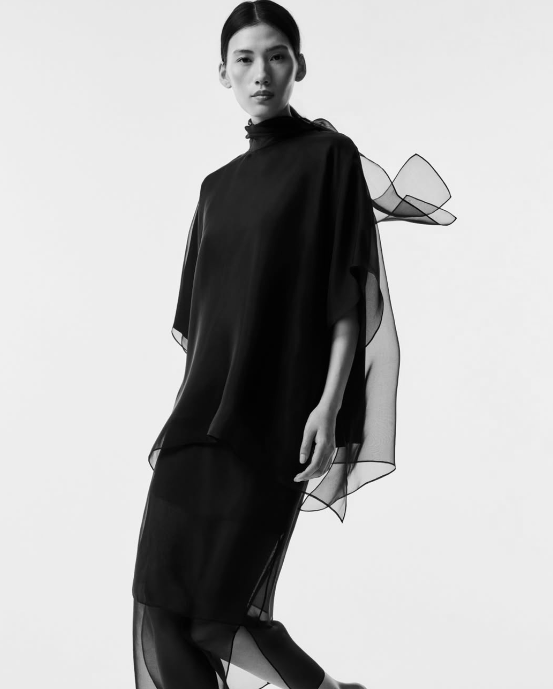 A model wearing a black high-neck, draped tunic-style dress with sheer overlay panels and a bow detail on the shoulder, photographed against a plain white studio background.