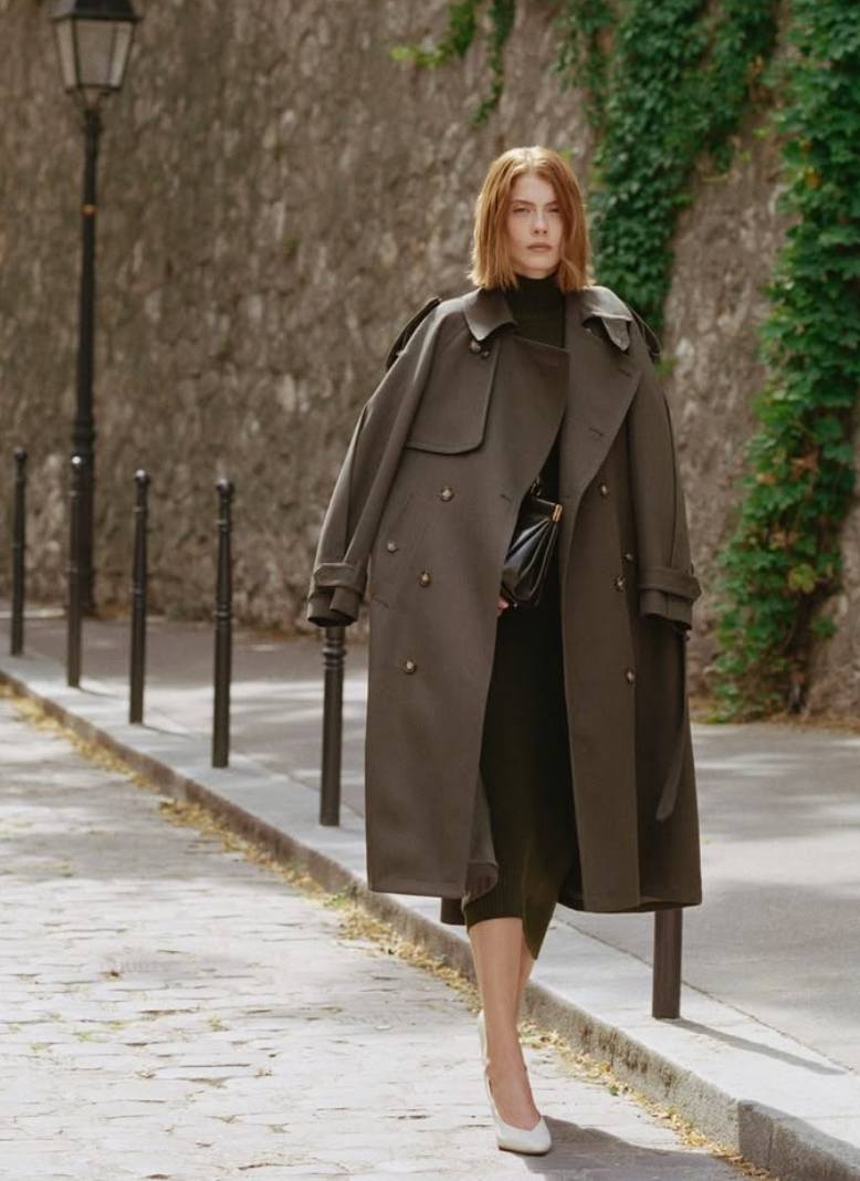 A woman walks down a cobblestone street wearing a long olive-brown trench coat over a dark outfit, with a stone wall and green ivy in the background.