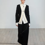 A model walks in a minimalist studio setting wearing a black and white fashion ensemble consisting of a structured black blazer with white contrast panels, a white shirt, and a long black skirt.