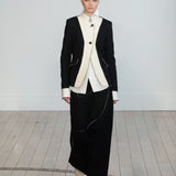 A model walks in a minimalist studio setting wearing a black and white fashion ensemble consisting of a structured black blazer with white contrast panels, a white shirt, and a long black skirt.