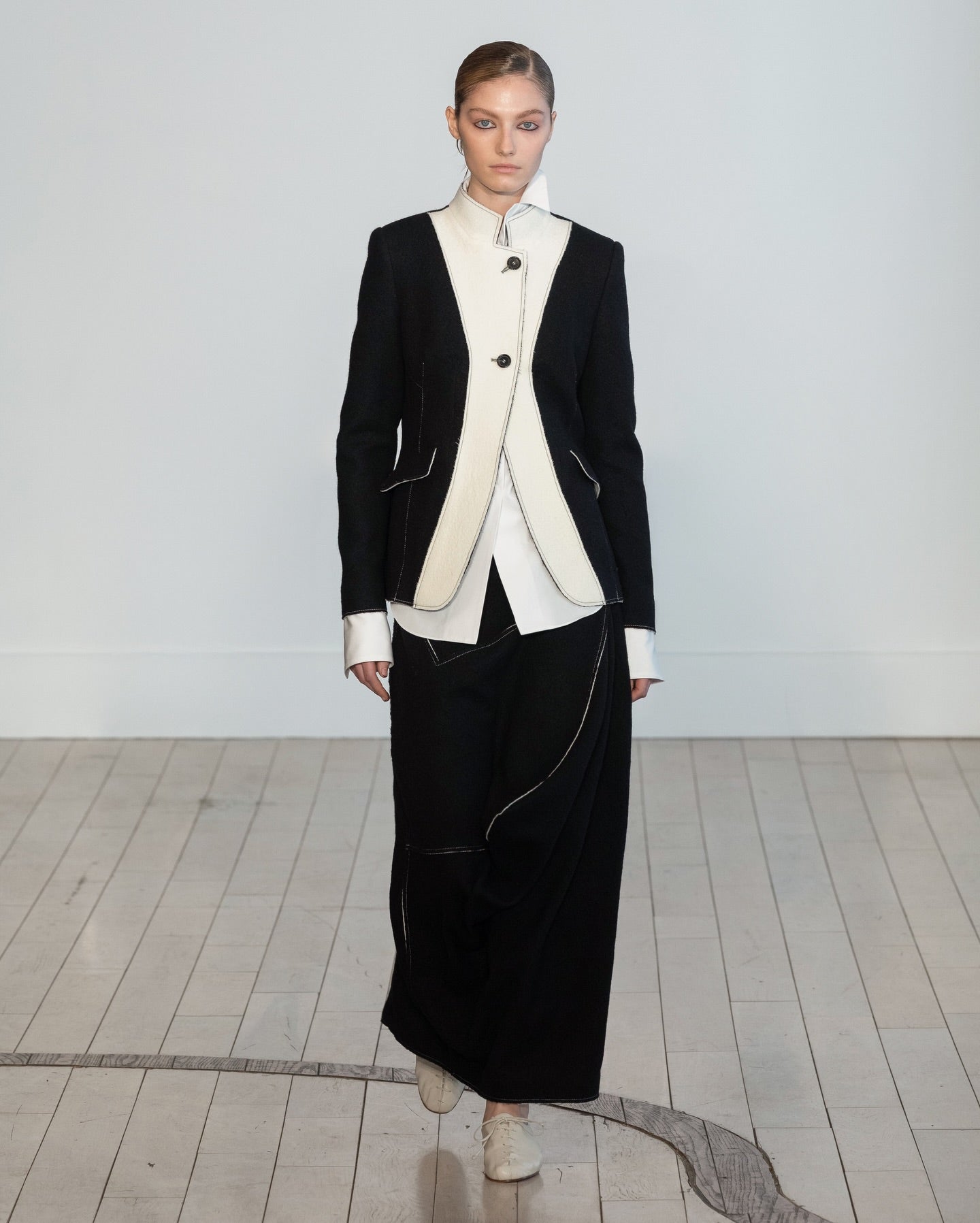A model walks in a minimalist studio setting wearing a black and white fashion ensemble consisting of a structured black blazer with white contrast panels, a white shirt, and a long black skirt.