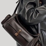 A dark brown leather shoulder bag from Gabriela Hearst with a front flap and gold-tone hardware, shown worn across the shoulder of a person wearing a black leather jacket against a light gray background.
