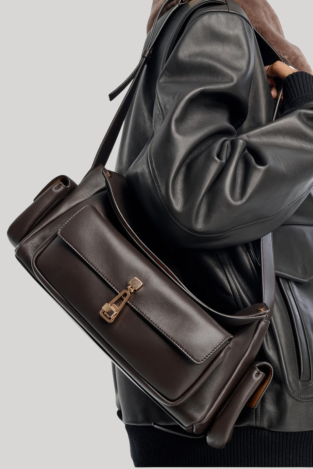 A dark brown leather shoulder bag from Gabriela Hearst with a front flap and gold-tone hardware, shown worn across the shoulder of a person wearing a black leather jacket against a light gray background.