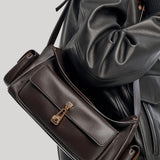 A dark brown leather shoulder bag from Gabriela Hearst with a front flap and gold-tone hardware, shown worn across the shoulder of a person wearing a black leather jacket against a light gray background.
