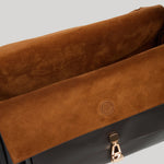 Close-up of a handbag from Gabriela Hearst showing a brown suede flap with a circular logo detail and a gold-tone zipper pull, set against a solid light gray background.