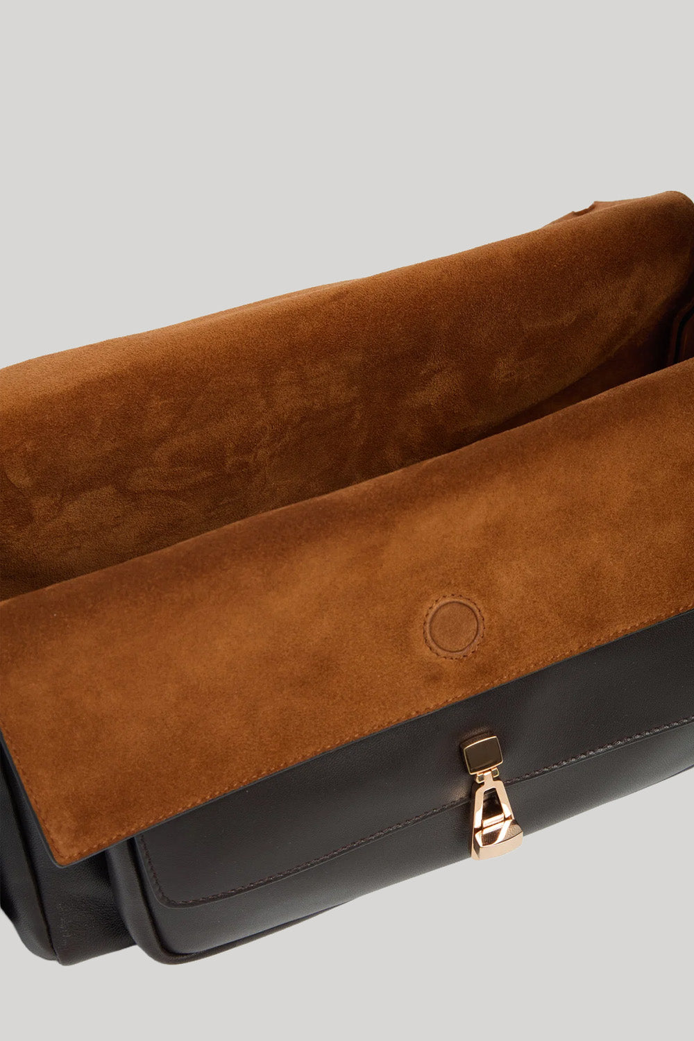 Close-up of a handbag from Gabriela Hearst showing a brown suede flap with a circular logo detail and a gold-tone zipper pull, set against a solid light gray background.