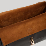 Close-up of a handbag from Gabriela Hearst showing a brown suede flap with a circular logo detail and a gold-tone zipper pull, set against a solid light gray background.