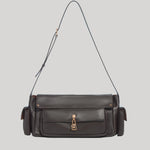 A dark brown leather shoulder bag from Gabriela Hearst with a front flap pocket, gold-tone hardware, and a thin matching leather strap, shown against a solid light gray background.