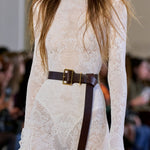 A close-up of a model wearing a cream lace long-sleeve mock-neck dress with a dark brown leather belt featuring a gold buckle, from Gabriela Hearst, set against a blurred runway background.