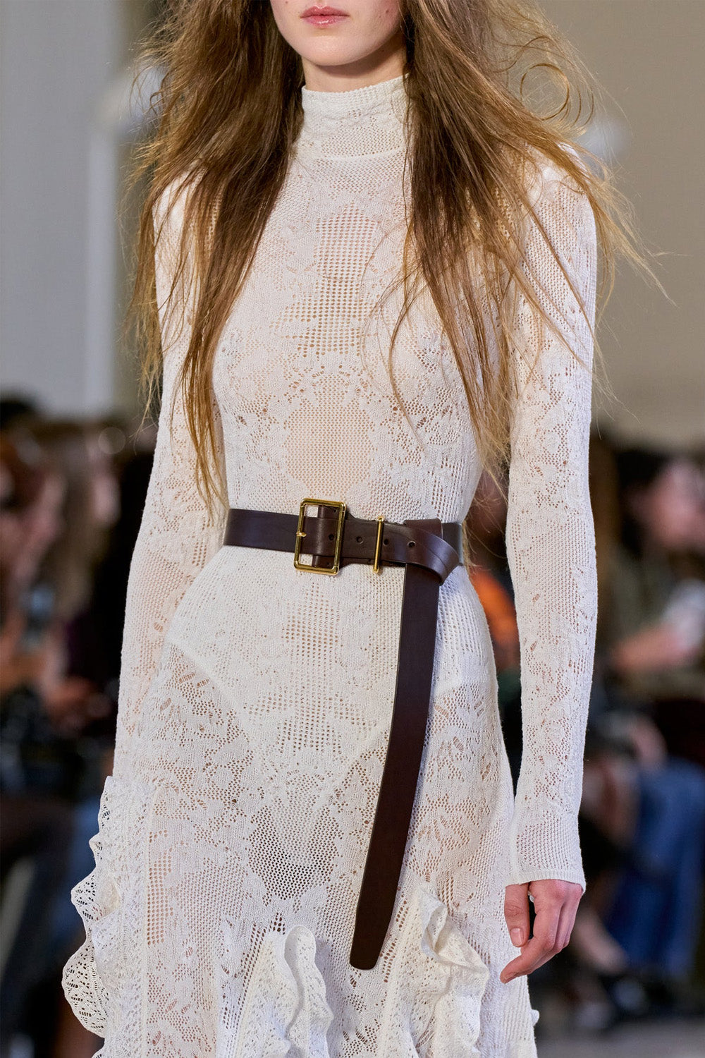 A close-up of a model wearing a cream lace long-sleeve mock-neck dress with a dark brown leather belt featuring a gold buckle, from Gabriela Hearst, set against a blurred runway background.