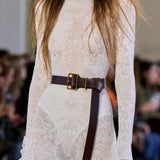 A close-up of a model wearing a cream lace long-sleeve mock-neck dress with a dark brown leather belt featuring a gold buckle, from Gabriela Hearst, set against a blurred runway background.