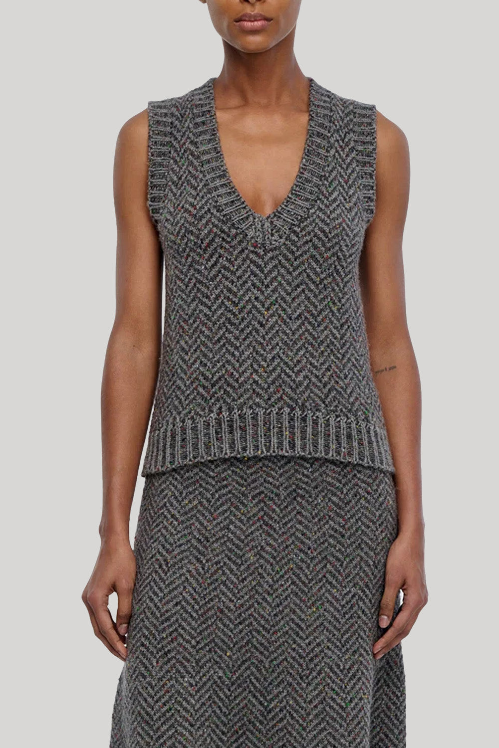 A model wearing a sleeveless knit vest and matching skirt in a gray and black herringbone pattern, from Gabriela Hearst shown from the neck down against a solid light gray background.
