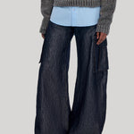 A model wearing dark wide-leg cargo pants with side flap pockets, a light blue button-up shirt, and a gray textured knit sweater, from Gabriela Hearst, shown from the chest down against a plain light gray background.