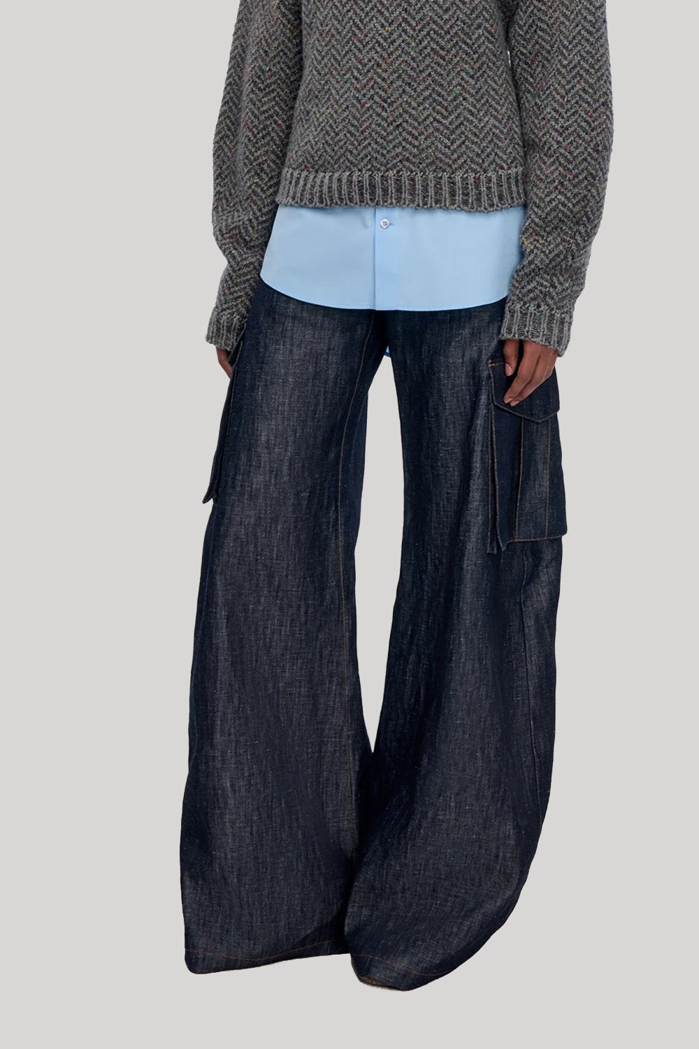 A model wearing dark wide-leg cargo pants with side flap pockets, a light blue button-up shirt, and a gray textured knit sweater, from Gabriela Hearst, shown from the chest down against a plain light gray background.