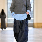 A model walks down a runway wearing a gray chevron turtleneck sweater, a light blue button-up shirt, loose dark blue denim cargo pants, and carrying a small black shoulder bag from Gabriela Hearst