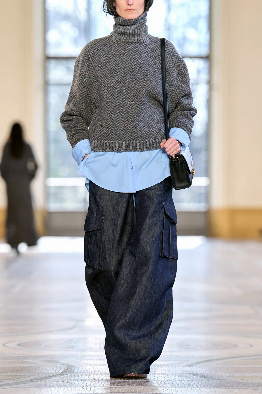 A model walks down a runway wearing a gray chevron turtleneck sweater, a light blue button-up shirt, loose dark blue denim cargo pants, and carrying a small black shoulder bag from Gabriela Hearst