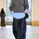 A model walks down a runway wearing a gray chevron turtleneck sweater, a light blue button-up shirt, loose dark blue denim cargo pants, and carrying a small black shoulder bag from Gabriela Hearst