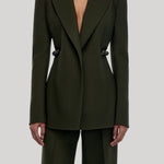 A model wearing a dark olive green tailored blazer with structured shoulders and matching wide-leg trousers, from Gabriela Hearst, shown against a light gray studio background.