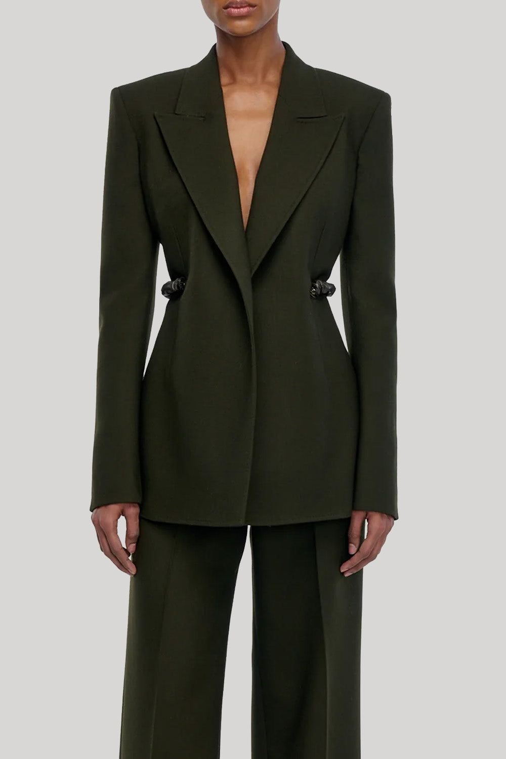 A model wearing a dark olive green tailored blazer with structured shoulders and matching wide-leg trousers, from Gabriela Hearst, shown against a light gray studio background.
