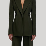 A model wearing a dark olive green tailored blazer with structured shoulders and matching wide-leg trousers, from Gabriela Hearst, shown against a light gray studio background.