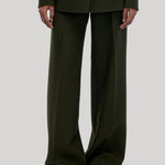 A model wearing dark olive green wide-leg trousers with a matching blazer and brown slippers, from Gabriela Hearst, shown from the shoulders down against a light gray studio background.