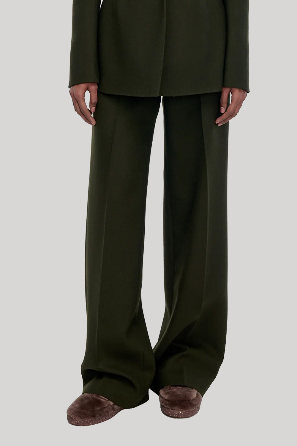 A model wearing dark olive green wide-leg trousers with a matching blazer and brown slippers, from Gabriela Hearst, shown from the shoulders down against a light gray studio background.