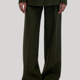 A model wearing dark olive green wide-leg trousers with a matching blazer and brown slippers, from Gabriela Hearst, shown from the shoulders down against a light gray studio background.