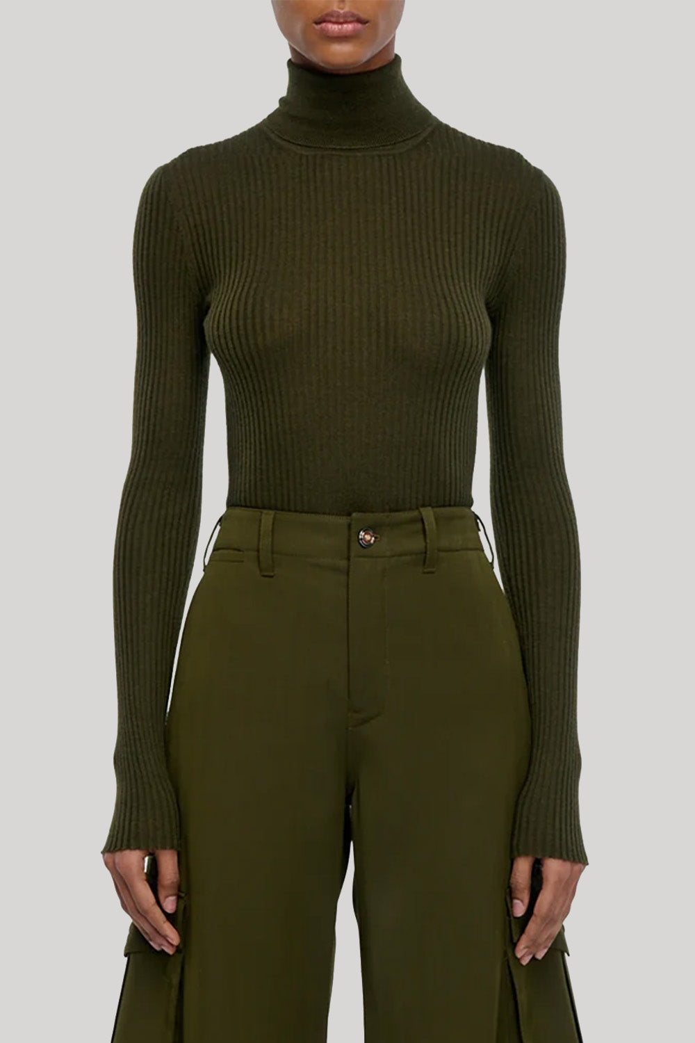 A model wearing a ribbed olive green mock neck long-sleeve top and matching olive green trousers, from Gabriela Hearst, shown from the mouth down against a solid light gray background.