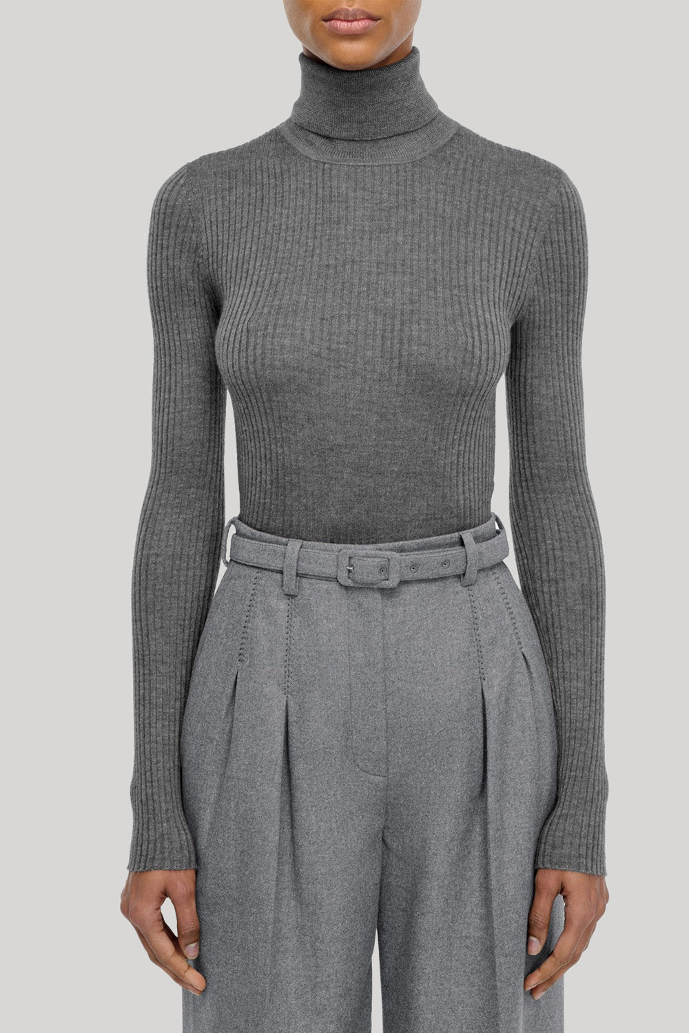 A model wearing a ribbed gray long-sleeve turtleneck sweater paired with high-waisted gray pleated trousers and a matching belt, from Gabriela Hearst, shown against a solid light gray background.