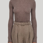 A model wearing a ribbed long-sleeve turtleneck sweater in taupe-brown, paired with high-waisted pleated trousers in a matching brown tone, from Gabriela Hearst, against a solid light gray background.