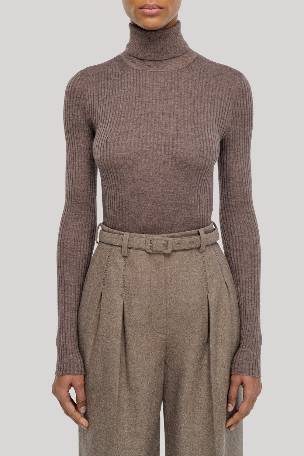 A model wearing a ribbed long-sleeve turtleneck sweater in taupe-brown, paired with high-waisted pleated trousers in a matching brown tone, from Gabriela Hearst, against a solid light gray background.