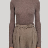 A model wearing a ribbed long-sleeve turtleneck sweater in taupe-brown, paired with high-waisted pleated trousers in a matching brown tone, from Gabriela Hearst, against a solid light gray background.