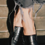 A close-up of a person wearing black leather penny loafers with frayed gray jeans, standing against a rustic wooden background.