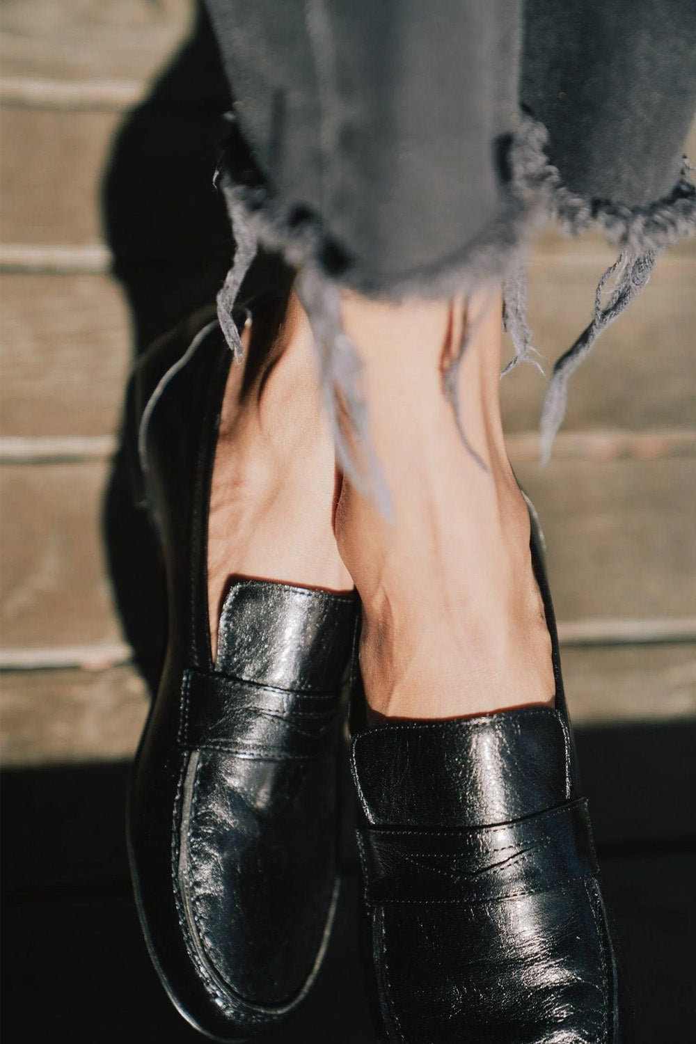 A close-up of a person wearing black leather penny loafers with frayed gray jeans, standing against a rustic wooden background.