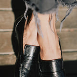 A close-up of a person wearing black leather penny loafers with frayed gray jeans, standing against a rustic wooden background.
