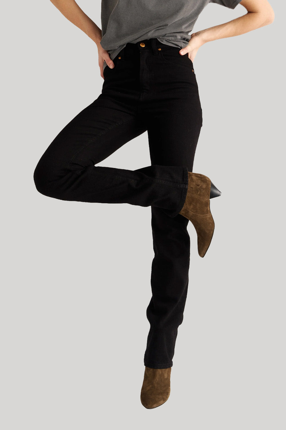 A model wearing black high-rise jeans, styled with a gray t-shirt and brown suede ankle boots, captured from the waist down against a solid light gray background.