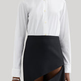 A model wearing a white long-sleeve Jil Sander button-up shirt tucked into a black asymmetrical wrap skirt with a diagonal slit, photographed against a neutral light gray background.