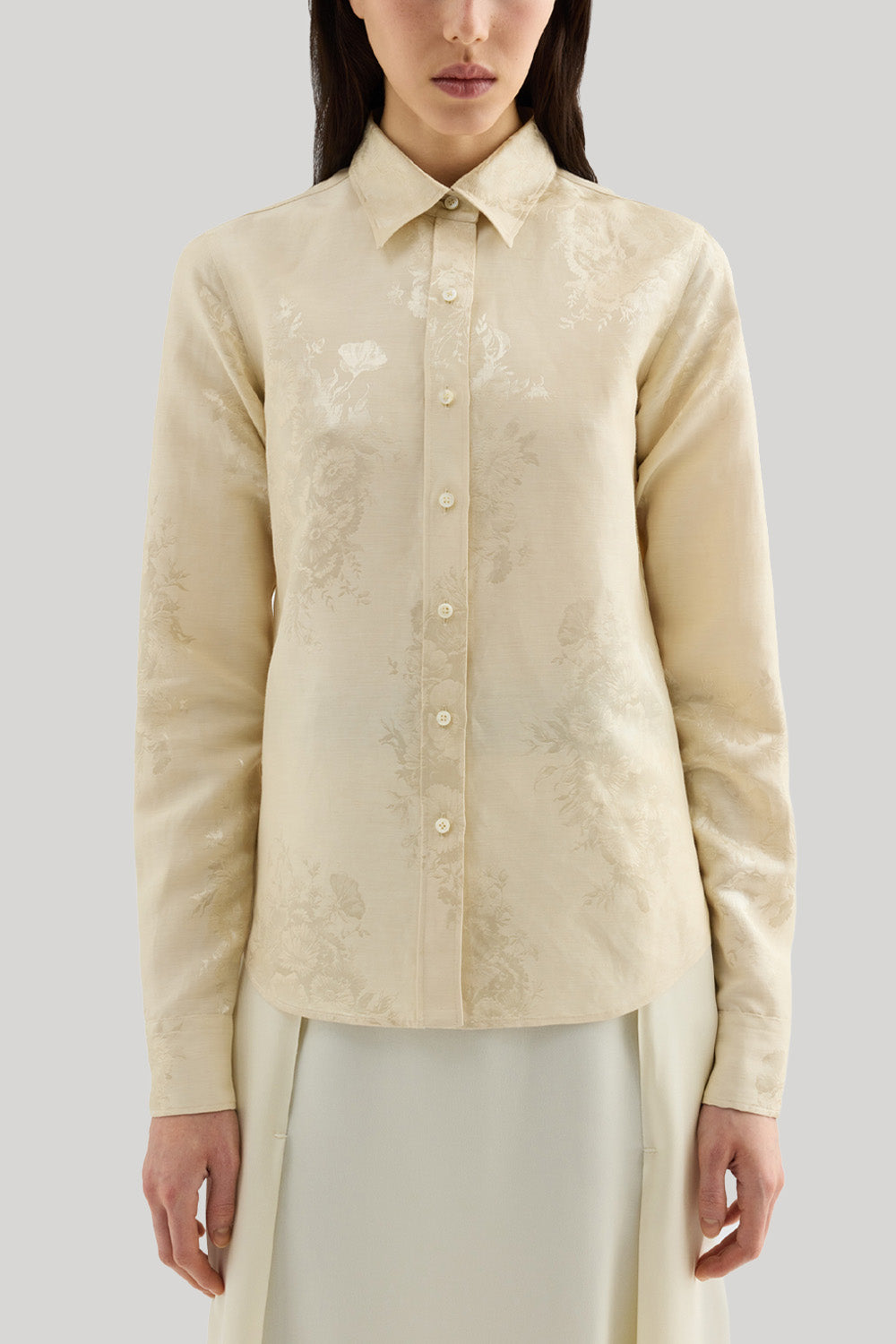 Front view of a woman wearing a cream-colored long-sleeve blouse with a subtle tonal floral jacquard pattern, paired with a matching cream skirt, against a solid light gray background. The shirt is Jil Sander 