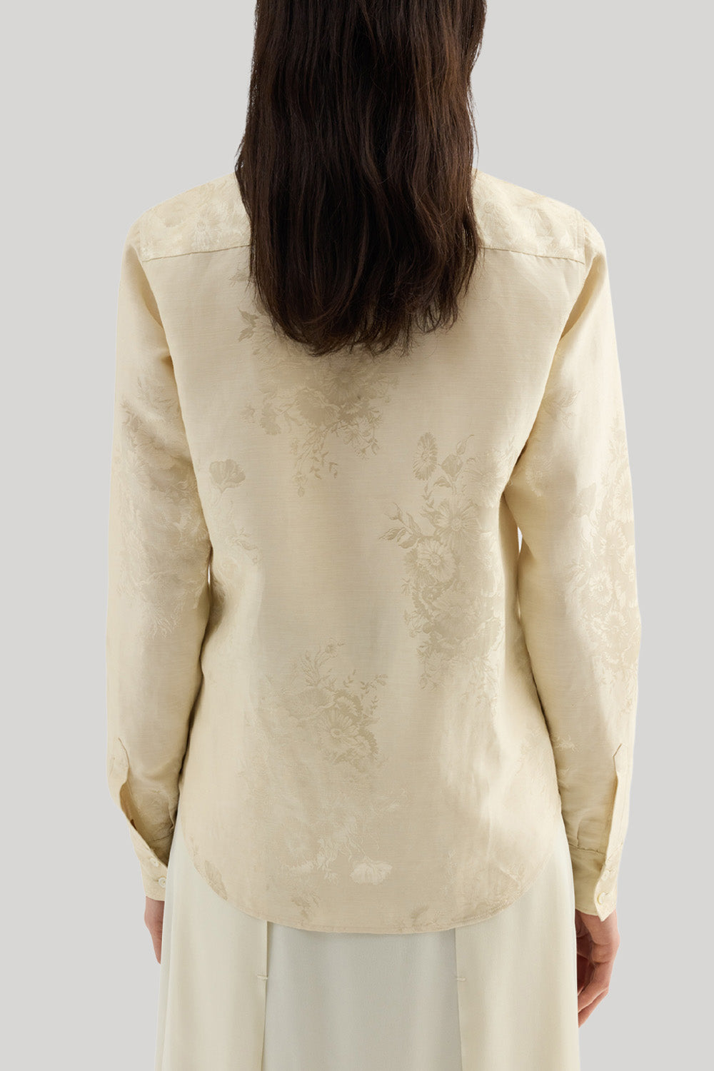 Back view of a woman wearing a cream-colored long-sleeve blouse with a subtle tonal floral jacquard pattern, paired with a matching cream skirt, against a solid light gray background. The shirt is Jil Sander 