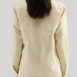 Back view of a woman wearing a cream-colored long-sleeve blouse with a subtle tonal floral jacquard pattern, paired with a matching cream skirt, against a solid light gray background. The shirt is Jil Sander 