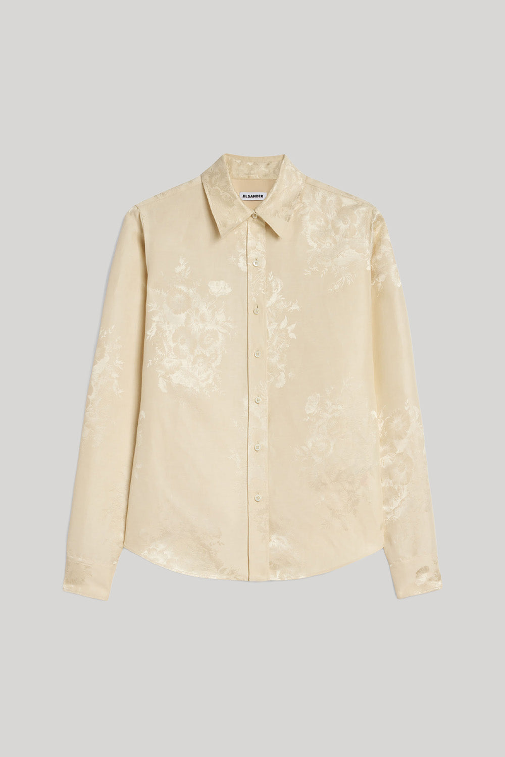 A light beige long-sleeve button-up shirt with a subtle tonal floral jacquard pattern from Jil Sander. Displayed flat against a neutral gray background.