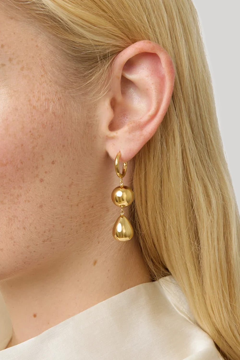 A close-up of a model's ear wearing a gold huggie hoop earring with two dangling polished gold beads.