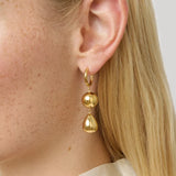 A close-up of a model's ear wearing a gold huggie hoop earring with two dangling polished gold beads.