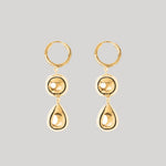 A pair of gold drop hoop earrings with a circular bead and teardrop pendant, shown against a solid light gray background.
