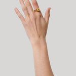 A gold signet ring with a polished, rounded drop worn on the pointer finger of a hand, shown against a solid light gray background.