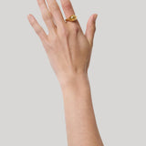 A gold signet ring with a polished, rounded drop worn on the pointer finger of a hand, shown against a solid light gray background.