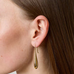 A close-up side profile of a woman wearing a delicate gold teardrop dangle earring, showing her ear, hair, and neck against a neutral light gray background.