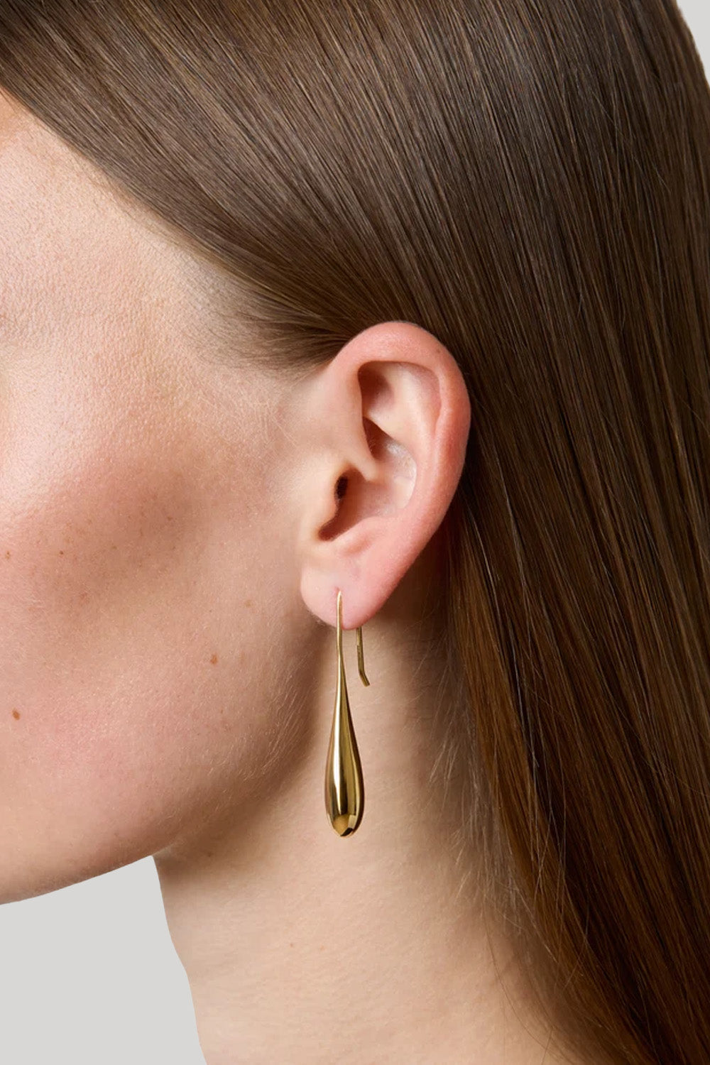 A close-up side profile of a woman wearing a delicate gold teardrop dangle earring, showing her ear, hair, and neck against a neutral light gray background.